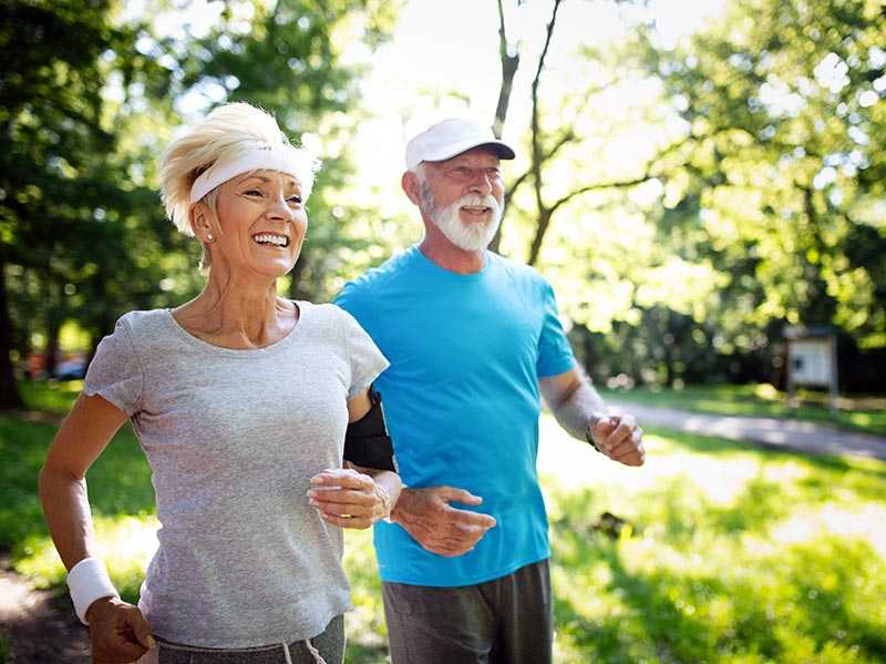 Älteres Paar lachend beim Jogging im sonnigen Park mit grünen Bäumen
